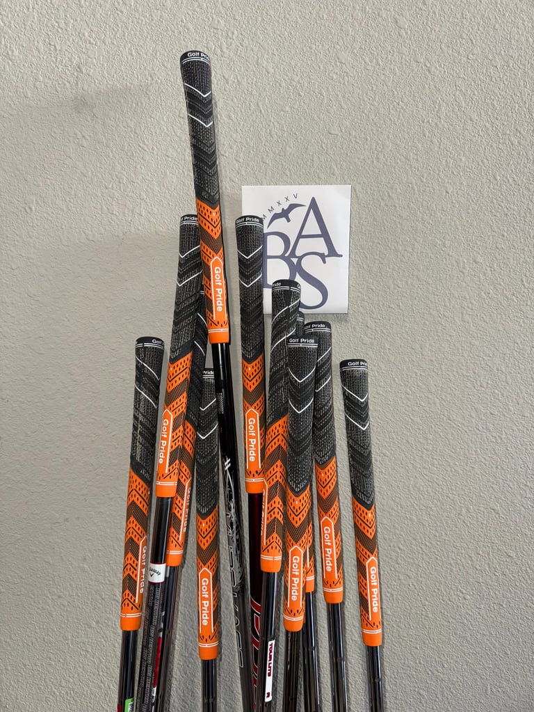 Collection of golf clubs with orange and black grips arranged vertically against a white wall with ABAS logo card
